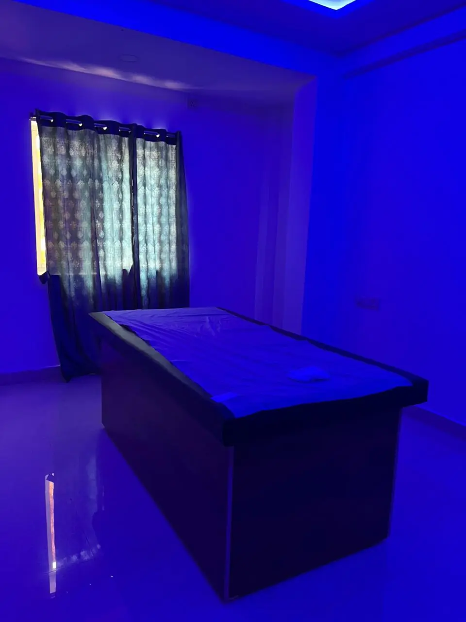 A treatment room at Sparkle Wellness Spa: a massage bed under soft blue light, beside a curtained window